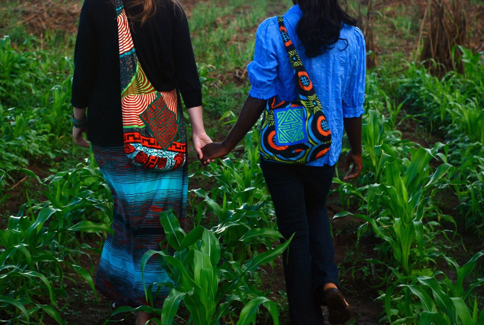 Two women holding hands as they walk through a green maize field, carrying patterned Nido shoulder bags